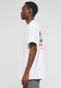 T-shirt en coton blanc à manches courtes, arborant un imprimé graphique au dos et un texte rouge. Le tissu semble doux et a une coupe décontractée.