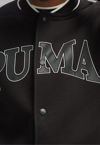 Veste universitaire noire à texture douce, avec un grand lettrage blanc "PUMA". Comprend des boutons-pression noirs et des accents de col rayés.