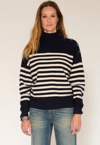 Femme aux cheveux longs portant un pull rayé bleu marine et blanc avec des boutons en forme d'ancre et un jean bleu, se tenant devant un fond uni.