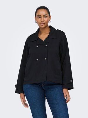 Kvinde iført sort dobbeltradet jakke med knappede manchetter og mørkeblå jeans, stående mod en ensfarvet lys baggrund.