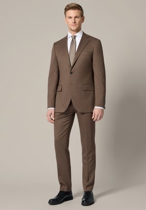 Mężczyzna stojący w dopasowanym brązowym garniturze z jednorzędową marynarką na jeden guzik, białą koszulą, wzorzystym brązowym krawatem i czarnymi eleganckimi butami na beżowym tle.