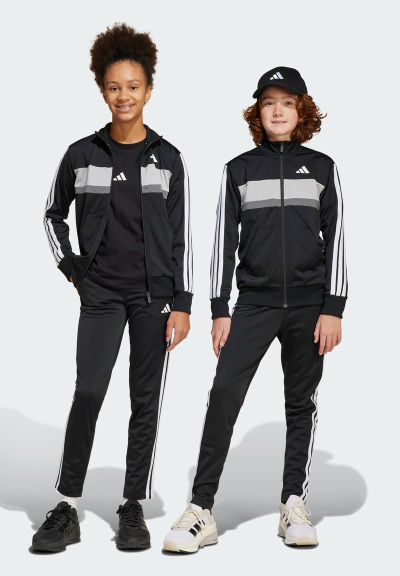 Deux individus en survêtements de sport noirs avec des rayures grises, des chaussures noires et des casquettes noires. Ils affichent le logo Adidas et trois rayures blanches le long des manches.