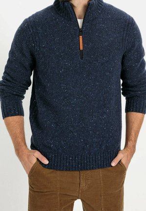 Hombre con un jersey de punto azul oscuro con cuello con cremallera hasta la mitad y pantalones de pana marrones, con las manos metidas en los bolsillos.