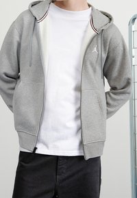 Sudadera gris con cremallera y dos bolsillos frontales, que cuenta con una capucha y detalles de rayas rojas y blancas en el cuello interior. Camiseta gráfica blanca debajo.