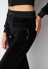 Schwarze Samtjogginghosen mit einer enganliegenden Passform, seitlichen Taschen, einem Kordelzug mit goldenen Akzenten und strukturierten Details an den Nähten.