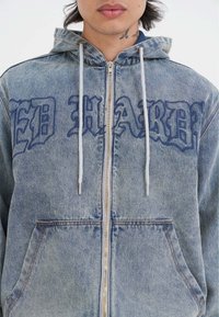 Sudadera de mezclilla con letras azules "ED HARDY" en el frente, cordones blancos y cremallera plateada. Cuenta con bolsillos frontales y una textura azul desgastada.