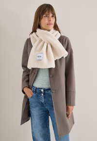 Faux fur creme tørklæde med label, løst viklet om halsen; parret med en grå top og en brun frakke, sammen med blå jeans.