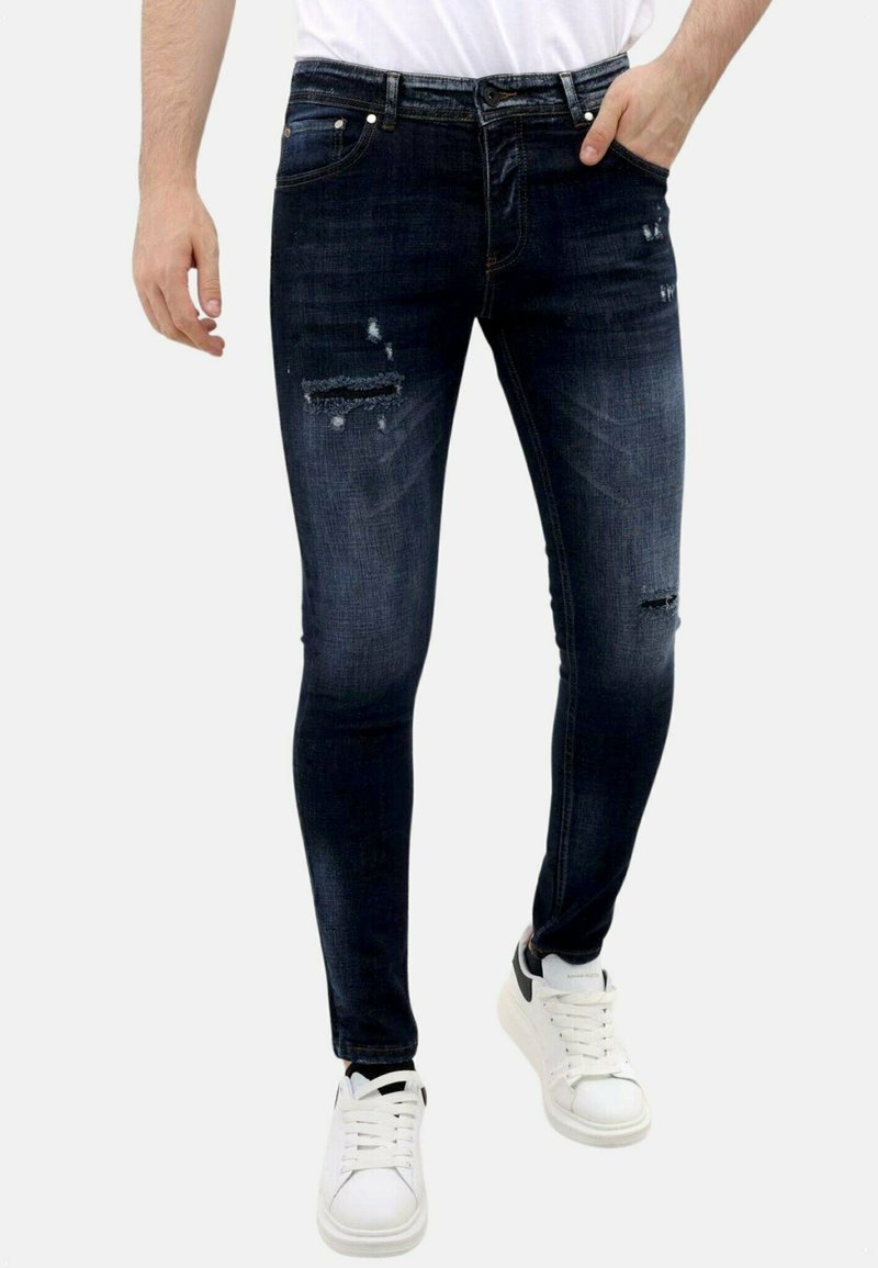 Donkerblauwe slim-fit jeans met versleten accenten en subtiele vervaging; heeft een standaard tailleband en voorzakken, gecombineerd met witte sneakers.