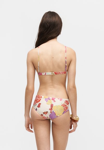 Femme aux longs cheveux bruns porte un bikini fleuri avec des fleurs roses, jaunes et rouges, debout dos à la caméra sur un fond uni.