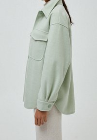 Camicia giacca oversized verde chiaro realizzata in tessuto morbido, dotata di colletto, una tasca frontale e polsini con bottoni.