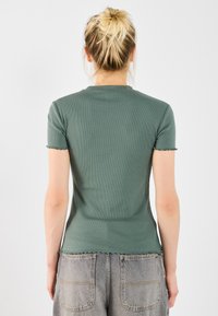 Green ribbed fitted t-shirt with short sleeves and frayed hem details, paired with gray high-waisted jeans, viewed from the back.