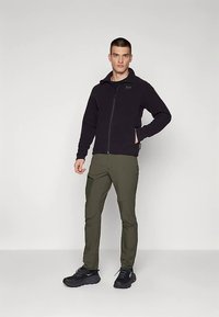 Svart fleecejacka med dragkedja, matchad med olivgröna byxor. Modellen står i mörka sportskor mot en neutral bakgrund.