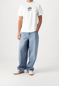 Biała bawełniana koszulka z graficznym logo, w połączeniu z luźnymi jasnoniebieskimi dżinsami i kremowymi sneakersami z czerwonymi akcentami.