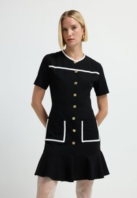 Donna che indossa un vestito nero a maniche corte con bottoni dorati, tasche con bordi bianchi e orlo arricciato, abbinato a collant bianchi con motivo.