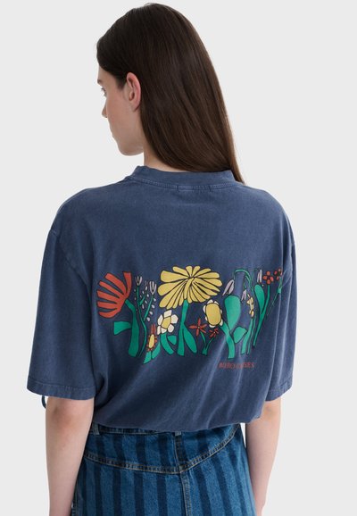 Femme aux longs cheveux bruns porte un t-shirt bleu oversize avec un motif floral abstrait coloré dans le dos, assorti à une jupe en denim rayée bleue.