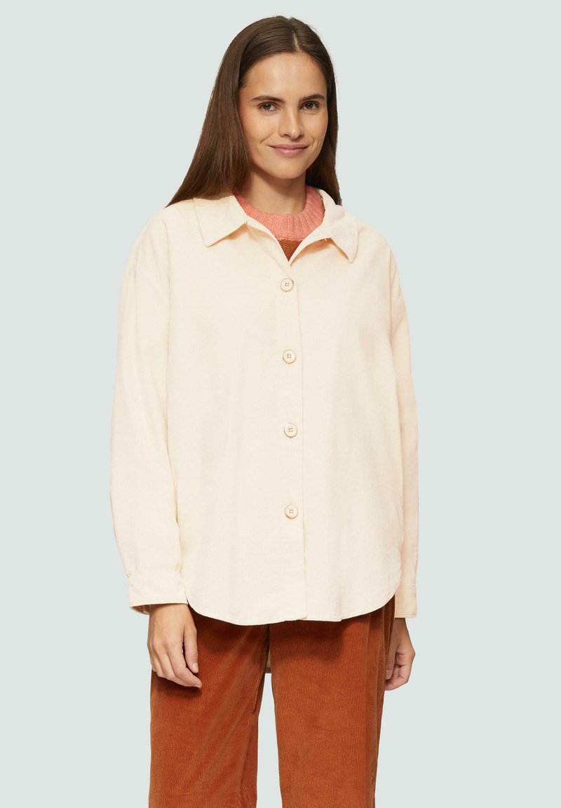 Licht perzikkleurig overhemd met knoopsluiting en kraag, enkele borstzak en lange mouwen. Zachte textuur. Draagt over een oranje gebreide trui.