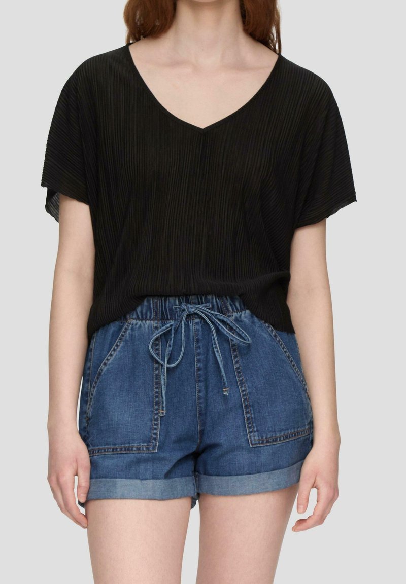 Femme portant un haut noir à manches courtes en V et un short en jean bleu avec ourlet roulé et taille à cordon, debout devant un fond uni.