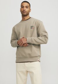 Beige sweatshirt made of cotton blend, features a round neckline, ribbed cuffs, and a small logo patch on the chest. Paired with light-colored pants.