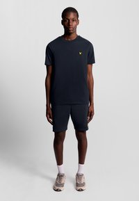 Donkerblauwe t-shirt en bijpassende shorts, beide van een katoenen mengsel. Heeft een klein geel logo op de borst. Model draagt lichtgekleurde sneakers.