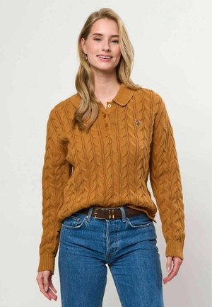 Junge Frau mit langen blonden Haaren, die einen braunen Zopfstrickpullover und blaue Jeans trägt, steht vor einem schlichten hellen Hintergrund.