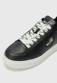 Zwarte leren sneaker met witte, gestructureerde veters, hartvormig logo en geperforeerde teendetail; heeft een witte rubberen zool.
