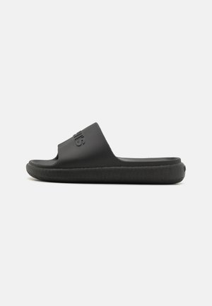 Chanclas negras de goma texturizada con una correa ancha y suela estriada, que presentan un logo en la parte superior.