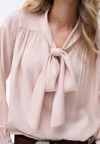 Jasnoróżowa bluzka z wiązaniem przy dekolcie, z miękkiego materiału, z plisowanymi detalami na ramionach i długimi rękawami, zestawiona z brązowym paskiem.