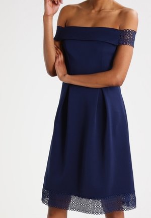 Kvinde iført en mørkeblå off-shoulder kjole med blondekant på ærmer og skørt, stående med den ene arm krydset og den anden hånd nær ansigtet.