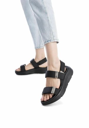 Sandalias deportivas negras con correas ajustables, detalles texturizados y suela gruesa. El diseño presenta acentos metálicos y una base plana.