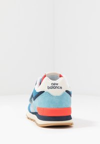 Sneaker en suede bleu et mesh avec des accents blancs et rouges, arborant une semelle en caoutchouc et le logo "New Balance" sur le talon.
