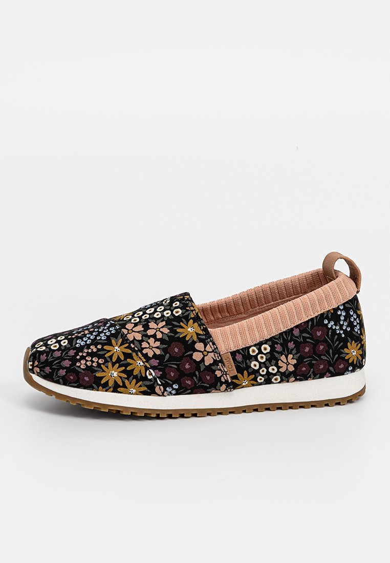 Zapato tipo slip-on con parte superior de tela negra que presenta un patrón floral multicolor, cuello acanalado rosa, suela blanca y suela de goma marrón.