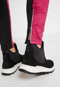 Botas deportivas negras con un upper texturizado negro, suela blanca y forro gris, combinadas con leggings ajustados en rosa y negro con puños con cremallera.