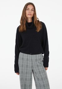 Woman with long brown hair wearing a black turtleneck sweater and gray plaid wide-leg pants standing against a white background.