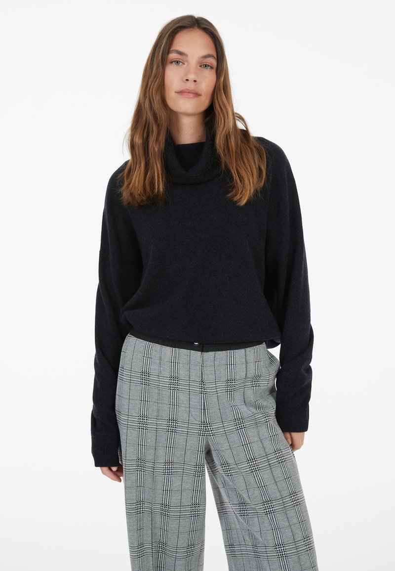 Woman with long brown hair wearing a black turtleneck sweater and gray plaid wide-leg pants standing against a white background.