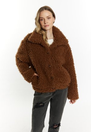 Jeune femme portant une veste marron duveteuse boutonnée avec col et un jean noir déchiré sur fond blanc.
