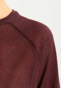 Pull en maille bordeaux avec une finition texturée, un col rond et des manches raglan. Le tissu semble léger et respirant.
