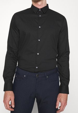 Homme portant une chemise noire cintrée boutonnée et un pantalon texturé bleu marine foncé, debout devant un fond uni.