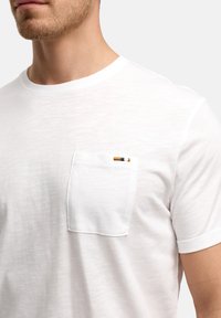 Bijela majica kratkih rukava od mekog materijala, s džepom na prsima koji ima žute i plave horizontalne pruge. Glatka tekstura, ležeran dizajn.