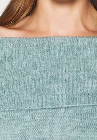 Světlomodrý žebrovaný pletený svetr se širokým výstřihem, s texturovaným vzorem a měkkým povrchem. Horní a dolní části jsou výrazně odlišné.