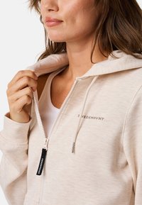 Light beige zip-up hoodie made of soft fabric, featuring a drawstring hood, white zipper, and subtle logo on the chest.