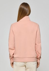 Rosa fleece sweatshirt med hög krage, ribbade ärmslut och nederkant. Slät textur och avslappnad passform, bärs över ljusa byxor.