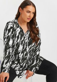 Camicia astratta in bianco e nero con colletto, con bottoni frontali, maniche lunghe e un tessuto testurizzato. Il modello è seduto.