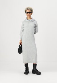 Vestido de malha cinza-claro com gola drapeada, mangas longas e punhos canelados, combinado com botas pretas de atacadores e uma bolsa preta.