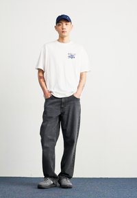 Bela bombažna majica s svetlo modrim grafičnim tiskom, v kombinaciji s temno sivimi širokimi kavbojkami in sivimi športnimi copati. Model nosi modro kapo.