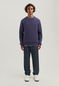 Paarse ribbel gebreide trui met een ronde hals, in combinatie met donkergrijze broek met wijde pijpen en witte sneakers. Eenvoudig, textuur design.
