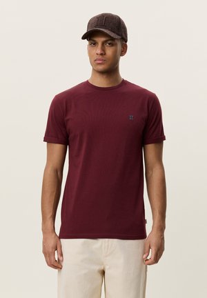 Homme portant un t-shirt bordeaux avec un petit logo bleu sur la poitrine et un pantalon beige, également coiffé d'une casquette de baseball marron texturée, debout devant un fond uni.