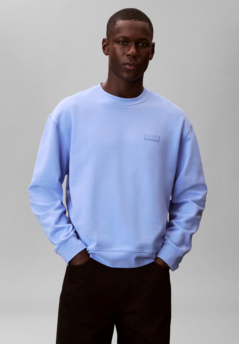 Jeune homme portant un sweat-shirt Calvin Klein bleu clair, les mains dans les poches, debout devant un fond gris uni.