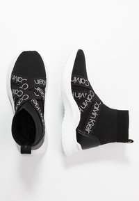 Černé pletené kotníkové tenisky s bílými podrážkami, které mají výrazné černé popruhy s logem "Calvin Klein". Hladká textury, moderní design.