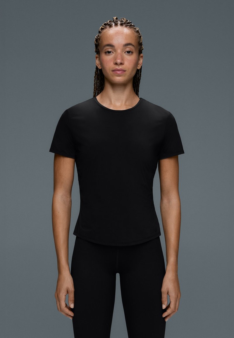 Jeune femme aux cheveux tressés portant un t-shirt noir moulant à manches courtes et un pantalon noir, debout devant un fond gris uni.