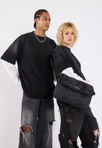 Black messenger bag with a textured finish, featuring adjustable straps and a flap closure. Worn with distressed black jeans and oversized black tees.
